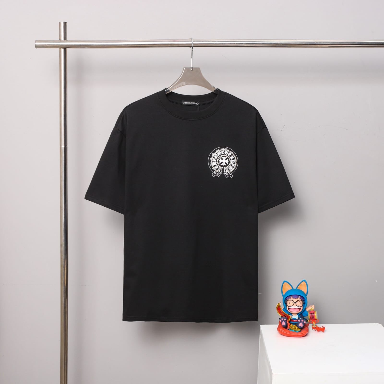 Black t-shirt with a logo on a hanger against a gray wall with a small figurine on a white surface.